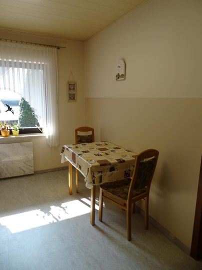 Een lichte kamer met een tafel en twee stoelen. Licht valt door het raam en er is een eenvoudige decoratie.