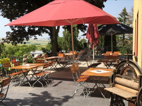 Eine einladende Terrasse mit mehreren Holzstühlen und Tischen unter roten Sonnenschirmen. Im Hintergrund sind grüne Bäume und eine weite Landschaft zu sehen.
