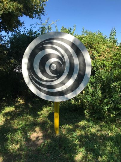 Ein großes Schild mit spiralförmigen schwarz-weißen Linien steht im Freien. Es ist von grünen Bäumen umgeben und zeigt einen klaren blauen Himmel.