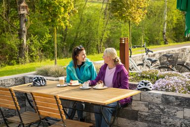 Zwei Frauen sitzen an einem Tisch im Freien und genießen Kaffee und Kuchen. Im Hintergrund sind Fahrräder und grüne Bäume zu sehen.