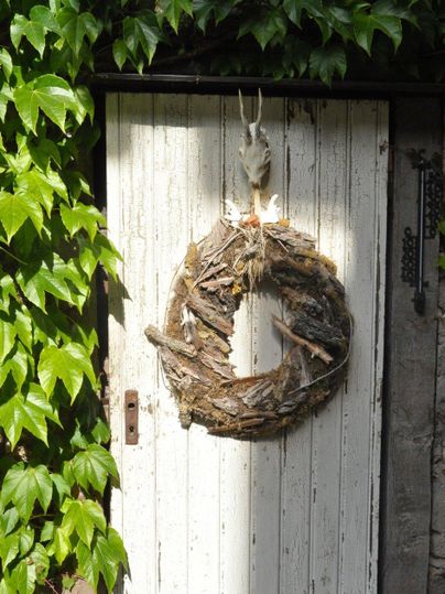 Ein rustikaler Kranz aus Ästen hängt an einer weißen Tür, die von grünem Efeu umgeben ist. Die Tür hat eine einfache, historische Ausstrahlung.