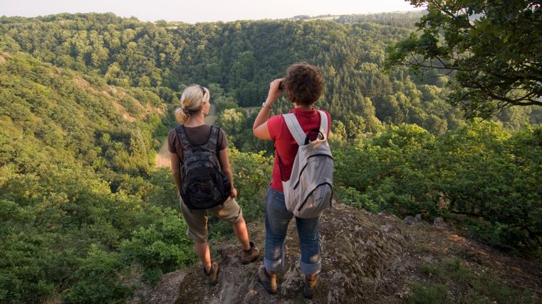 Zwei Frauen mit Rucksäcken stehen auf einem Felsen und blicken in ein bewaldetes Tal.