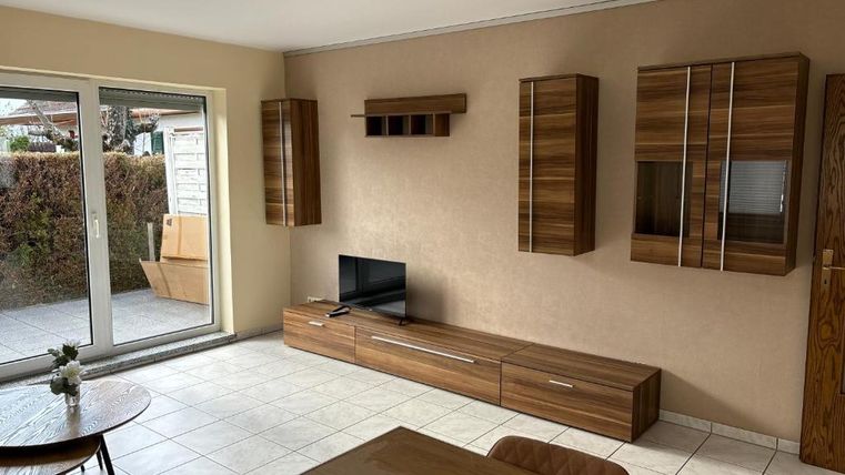 Ein modernes Wohnzimmer mit einem Fernseher und Holzmöbeln. Große Fenster sorgen für viel Tageslicht und einen Blick auf den Garten.