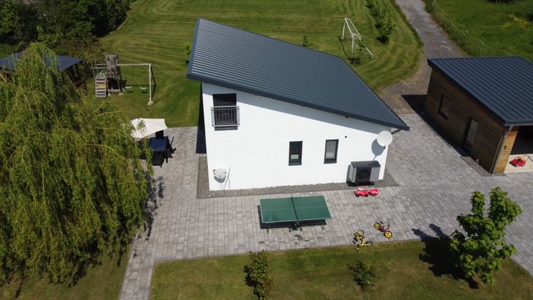 Une maison moderne blanche avec un toit gris se dresse sur une cour pavée. En arrière-plan, on peut voir des équipements de jeu et un espace vert bien entretenu.