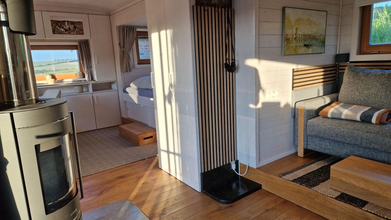 Ein heller, moderner Wohnraum mit Holzfußboden und einer gemütlichen Couch. Im Hintergrund sind Fenster mit Blick ins Freie und ein Kamin zu sehen.