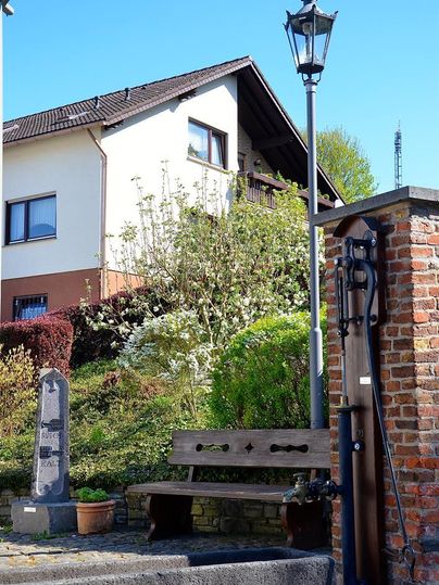 Eine gemütliche Ecke mit einer Bank und einem Brunnen vor einem Haus. Es blühen Pflanzen und die Sonne scheint.