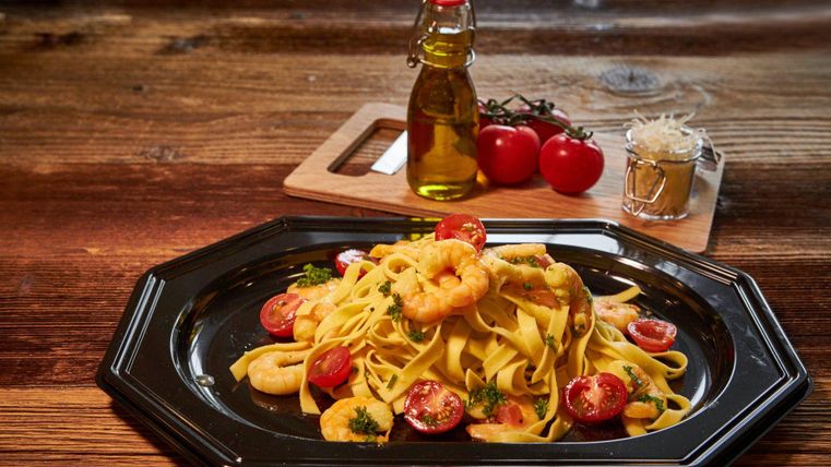 Ein Teller mit Tagliatelle, Garnelen und Cherry-Tomaten, garniert mit frischen Kräutern. Im Hintergrund stehen Olivenöl und eine Schale mit Parmesan.