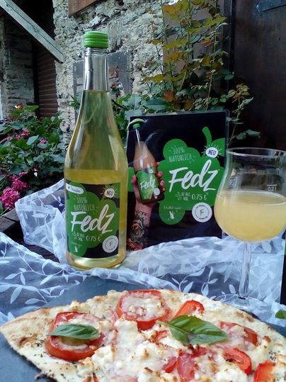 Ein Flaschengetränk namens Fedz steht neben einer Glasflasche und einem Karton in einem Garten. Im Vordergrund liegt eine Pizza mit Tomaten und frischem Basilikum.