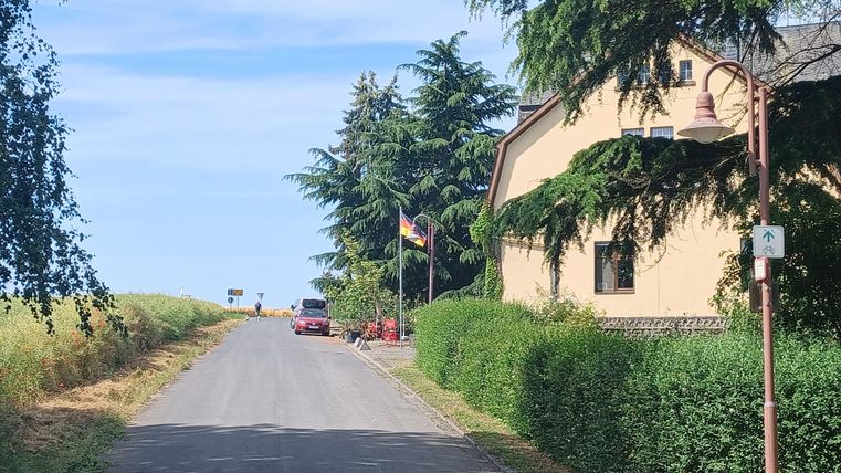 Eine ruhige Straße mit einem Haus an der Seite. Im Hintergrund sind Bäume und eine Fahne zu sehen.