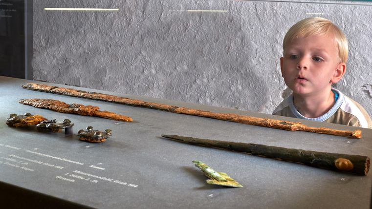 Ein neugieriger Junge schaut auf historische Ausstellungsstücke in einem Museum. Auf dem Tisch liegen verschiedene alte Gegenstände, darunter Stäbe und kleine Figuren.