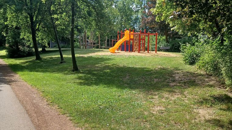 Ein Spielplatz mit einer Rutsche in einem grünen Park. Rundherum stehen Bäume und ein Spazierweg ist zu sehen.