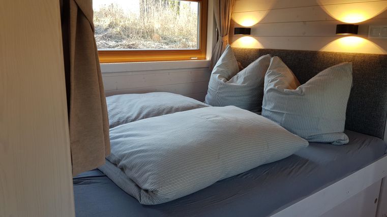Ein gemütliches Schlafzimmer mit einem großen Fenster und sanftem Licht. Auf dem Bett liegen große, einladende Kissen und Decken.
