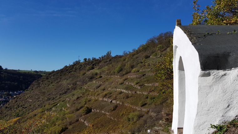 Ein weißes Gebäude in der Natur mit terrassierten Hügeln im Hintergrund. Der Himmel ist klar und blau.