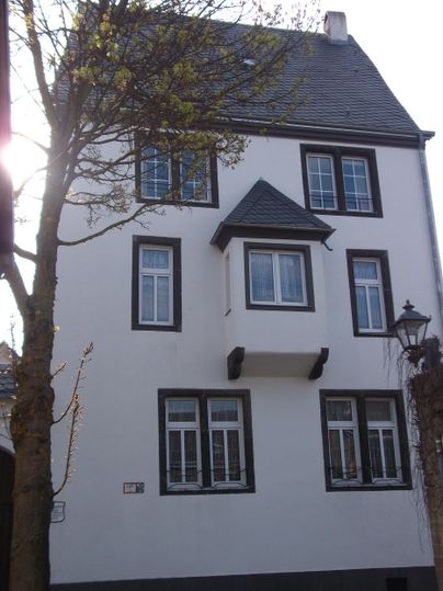 Ein dreigeschossiges, weißes Gebäude mit einem spitzen Dach. Es hat mehrere Fenster und steht neben einem Baum.