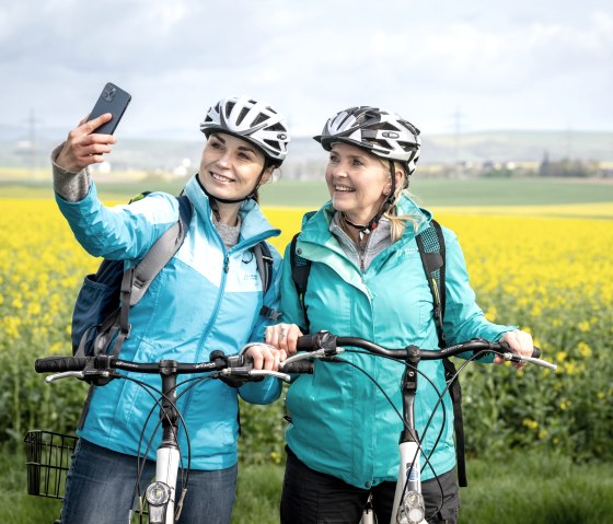 Zwei Frauen in blauen Jacken und Fahrradhelmen machen ein Selfie vor einem gelben Rapsfeld auf dem Maifeld-Radwanderweg., © Eifel Tourismus GmbH, Dominik Ketz