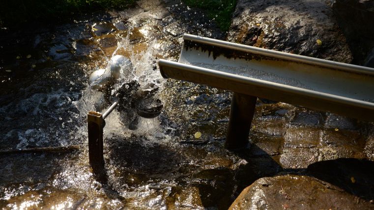 Un cours d'eau s'écoule sur un tas de pierres et est dirigé par un tube en métal. L'environnement est vert et ensoleillé.