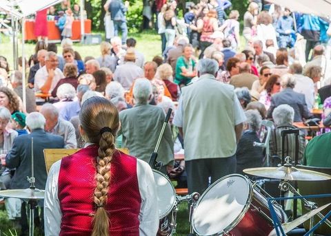 Bunte Wimpel hängen an einer Schnur vor einem klaren Himmel. Im Hintergrund sind grüne Bäume sichtbar.