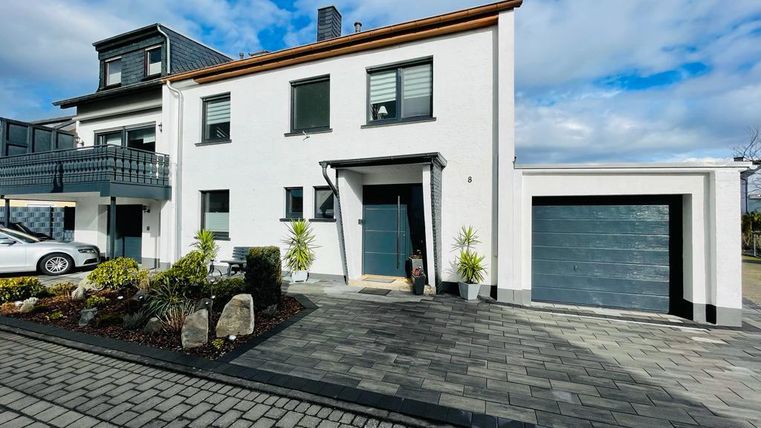 Ein modernes Haus mit weißen Wänden und grünen Fenstern. Der Vorgarten ist gepflegt und hat schöne Pflanzen.