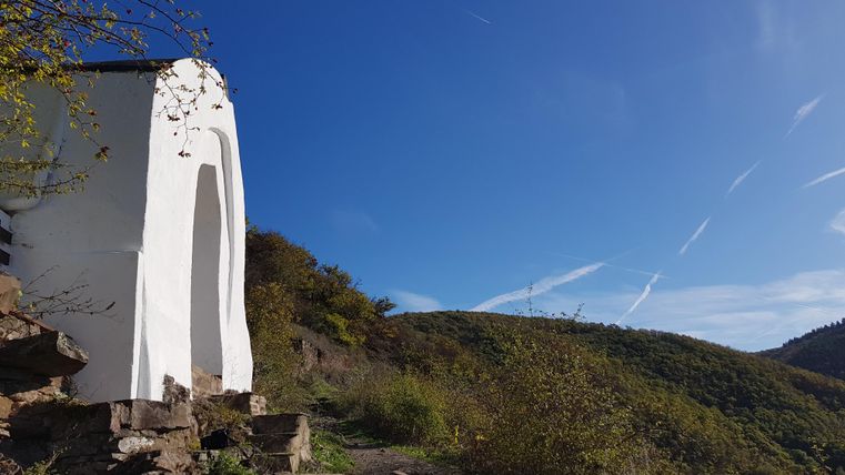 Eine weiße Kapelle steht auf einem Hügel mit grünen Bergen im Hintergrund. Der Himmel ist klar und blau.