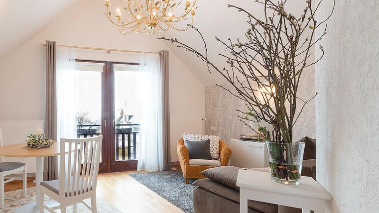 Ein modernes Wohnzimmer mit einer gemütlichen Sitzecke und einem eleganten Kronleuchter. Große Fenster sorgen für viel Licht und einen Blick nach draußen.