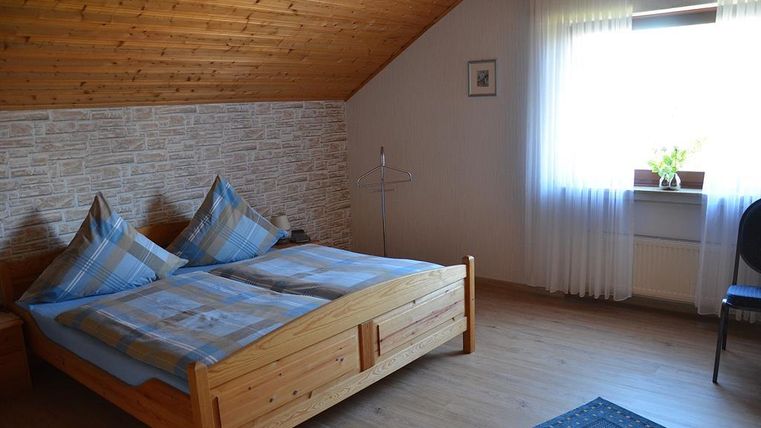 Ein gemütliches Schlafzimmer mit einem Holzbett und blauen Kissen. Es gibt ein Fenster mit Licht und einen Stuhl in der Ecke.