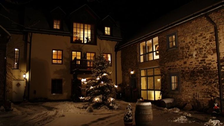 Ein gemütlicher Innenhof bei Nacht, umgeben von alten Steinhäusern. Ein Weihnachtsbaum ist festlich erleuchtet und der Boden ist mit Schnee bedeckt.