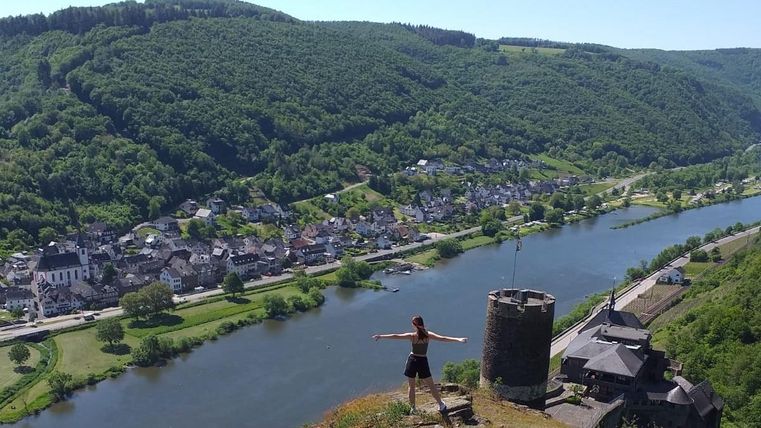 Eine Person steht auf einem Hügel mit weitreichendem Blick auf ein Dorf und den Fluss. Umgeben von grünen Hügeln bietet die Szene eine ruhige, natürliche Schönheit.
