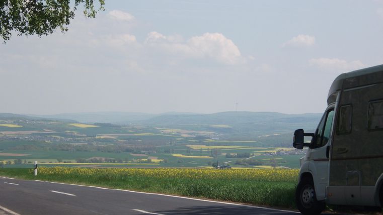 Ein Wohnmobil steht am Straßenrand mit Blick auf grüne Hügel und ein weitläufiges Feld. Der Himmel ist hell und wolkig.