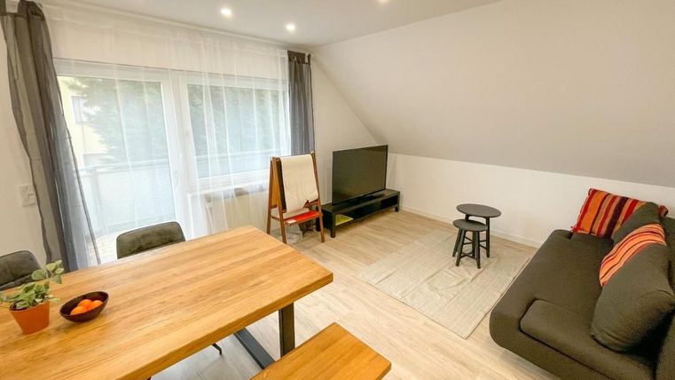 Ein modernes Wohnzimmer mit einem Esstisch, einem Sofa und einem Fernseher. Große Fenster lassen viel Licht herein und sorgen für eine helle, einladende Atmosphäre.