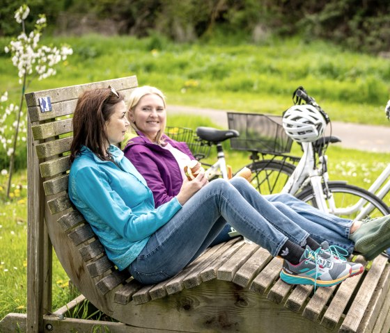 Zwei Frauen sitzen entspannt auf einer Holzbank im Grünen, mit Fahrrädern und Helmen daneben. Sie genießen eine Pause auf dem Maifeld-Radwanderweg., © Eifel Tourismus GmbH, Dominik Ketz