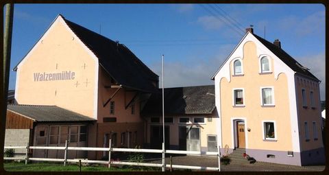 Ein altes Gebäude mit dem Schriftzug "Walzermühle" steht neben einem hellrosa Wohnhaus. Der Himmel ist blau mit wenigen Wolken.