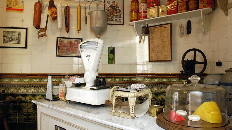 Een oude keukeninrichting met een weegschaal en verschillende voedingsmiddelen. Op de achtergrond hangen vleeswaren en blikken aan de muur.