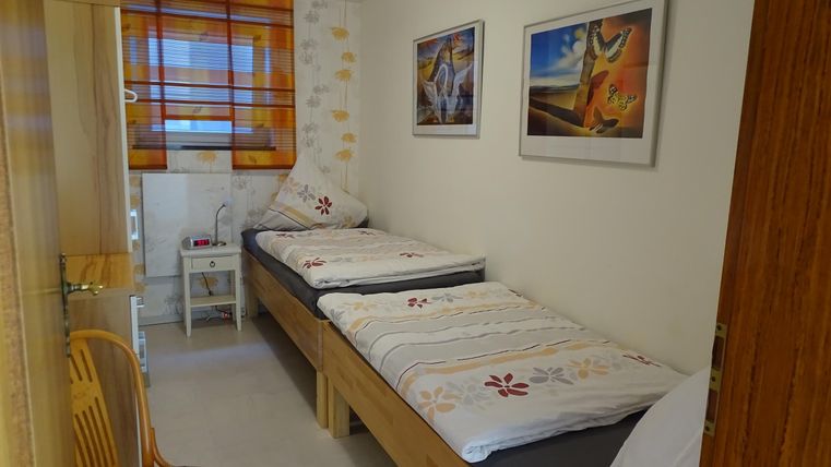 Een eenvoudige slaapkamer met twee eenpersoonsbedden en kleurrijke foto's aan de muur. Een stoel staat naast een klein tafeltje in de kamer.