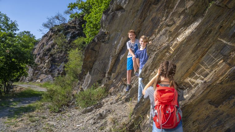 Zwei Kinder klettern an einem Felsen, während eine Person mit rotem Rucksack ein Foto macht.
