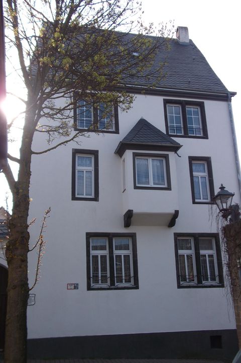Ein dreigeschossiges, weißes Gebäude mit einem spitzen Dach. Es hat mehrere Fenster und steht neben einem Baum.