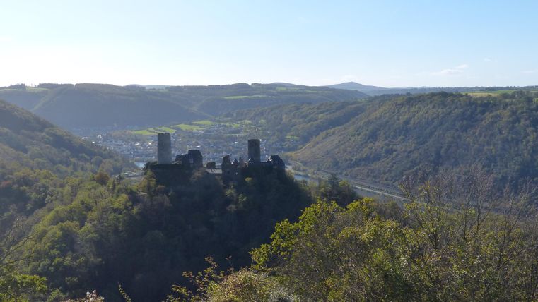 Eine alte Burg auf einem Hügel mit grünen Wäldern und einem Blick ins Tal. Im Hintergrund sind sanfte Hügel und ein klarer Himmel zu sehen.