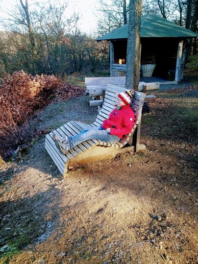 Eine Holzliege steht in der Natur, umgeben von Bäumen und Sträuchern. Eine Person in roter Kleidung sitzt entspannt auf der Liege.