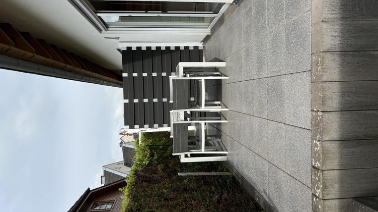 Eine Terrasse mit zwei weißen Stühlen und einem Tisch. Im Hintergrund ist ein Haus und eine Hecke sichtbar.