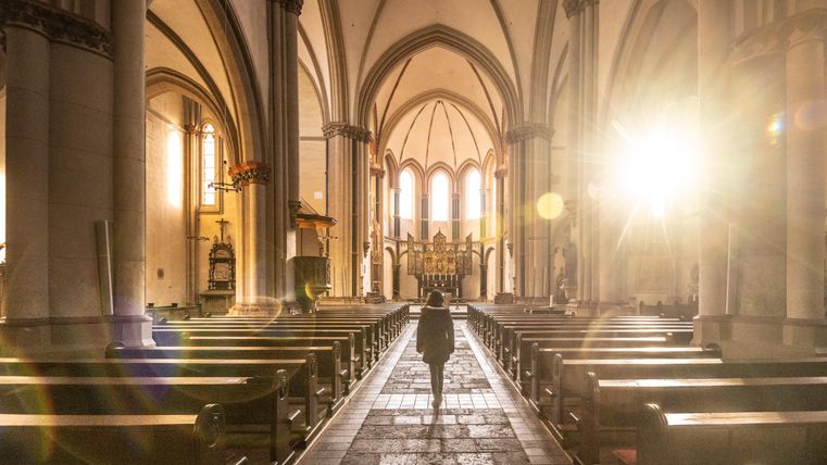 Ein lichtdurchfluteter Innenraum einer Kirche mit hohen Gewölben. In der Mitte steht eine Person zwischen den leeren Bankreihen.