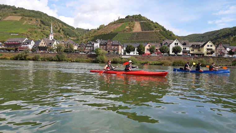 Zwei Kajakfahrer paddeln auf einem ruhigen Fluss. Im Hintergrund sind malerische Dörfer und Weinberge zu sehen.