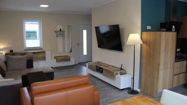 Un salon moderne avec un canapé confortable, une télévision au mur et un coin repas. Les couleurs chaudes et les meubles en bois donnent une atmosphère accueillante à la pièce.