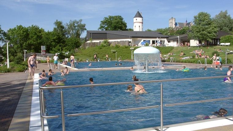 Ein Schwimmbad mit vielen Menschen, die im Wasser planschen. Im Hintergrund sind Bäume und Gebäude zu sehen.