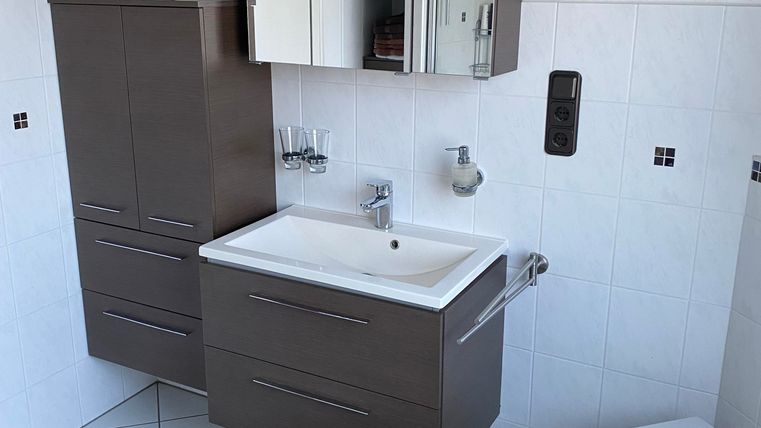 Ein modernes Badezimmer mit einem Waschbecken, Spiegel und einem WC. Der Boden ist mit weißen Fliesen und einem runden Teppich in Brauntönen ausgestattet.