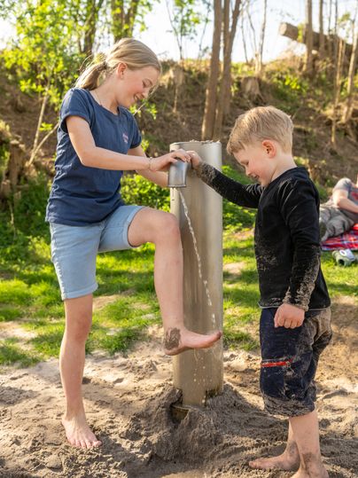 zwei Kinder säubern sich an einer Pumpe mit Wasser ihre Füße