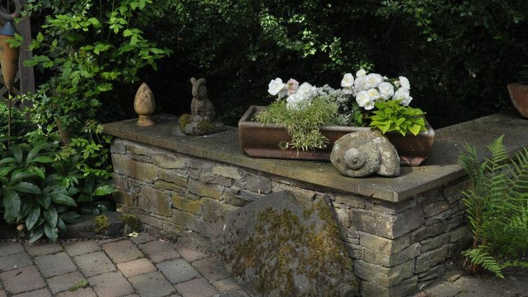 Ein schöner Steinschrank mit einer Pflanzenbox und weißen Blumen. Umgeben von grünen Pflanzen und kleinen Steinskulpturen.