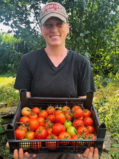 Eine Frau hält eine Kiste mit frischen Tomaten. Im Hintergrund sind Pflanzen und Bäume sichtbar.