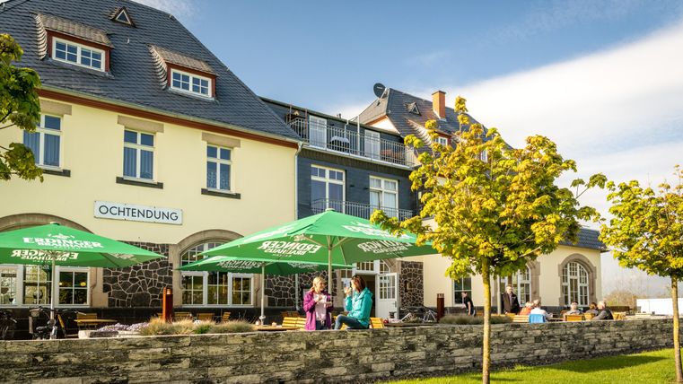 Ein charmantes Gebäude mit einer Terrasse und grünen Sonnenschirmen. Die Umgebung ist freundlich mit Bäumen und Rasenflächen.