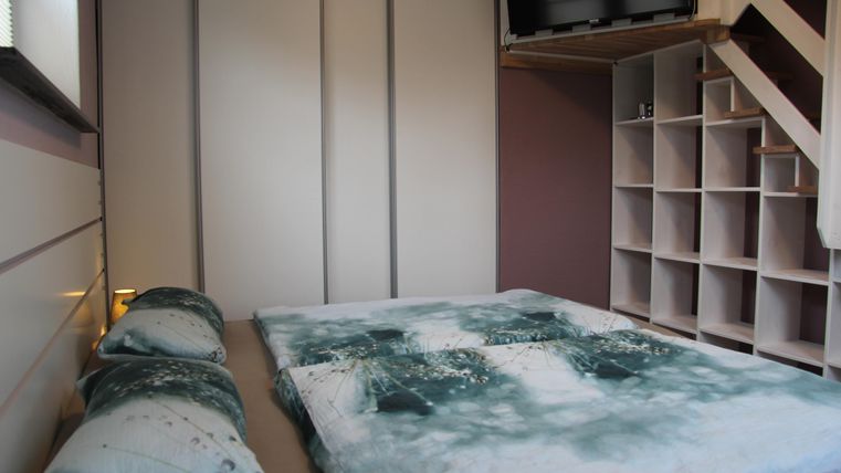 Une chambre moderne avec un futon et des coussins aux couleurs douces. En arrière-plan, des étagères et une télévision sont visibles.