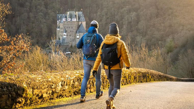 Zwei Wanderer gehen auf einem Weg, mit einem Schloss im Hintergrund. Die Sonne scheint in die Landschaft und schafft eine malerische Atmosphäre.
