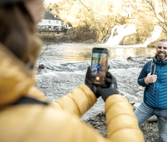 Ein Mann in blauer Jacke posiert l&auml;chelnd am Fluss, w&auml;hrend eine Person ihn mit einem Smartphone fotografiert. Im Hintergrund ein Wasserfall und ein Haus., &copy; Eifel Tourismus GmbH, D. Ketz